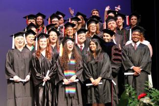 Four rows of smiling students in cap and gown, standing on the Fine Arts stage
