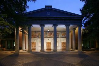 Fine Arts Building at twilight. Building has columns and a lighted porch with three wooden doors. A mural by Jean Charlot is above the doors.