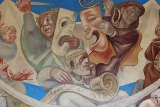 Photo of a mural featuring comedy and tragedy masks, held by players who are holding a knife and rope. A seated figure writes on a scroll, which reads "Athens the Beautiful" in Greek
