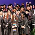 Four rows of smiling students in cap and gown, standing on the Fine Arts stage