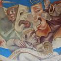 Photo of a mural featuring comedy and tragedy masks, held by players who are holding a knife and rope. A seated figure writes on a scroll, which reads "Athens the Beautiful" in Greek