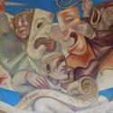 Photo of a mural featuring comedy and tragedy masks, held by players who are holding a knife and rope. A seated figure writes on a scroll, which reads "Athens the Beautiful" in Greek